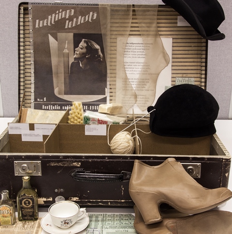 Musta salkku, jonka sisällä hattuja, lankarullia, mäntysuopaa ja edustalla korkokengät, kahvikuppi ja tassi, pieniä pulloja.