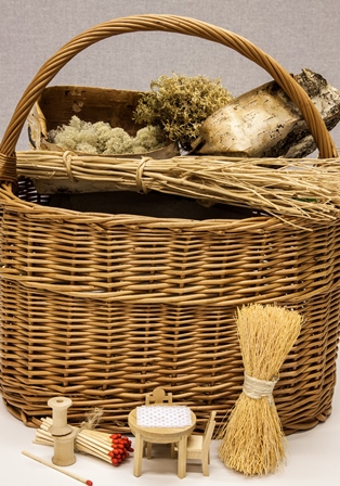 Punottu kori, josta pilkistää sammalta, jäkälää ja tuohta. Pöydällä korin vieressä pienoishuonekaluja, tulitikkuja ja lankarullia.