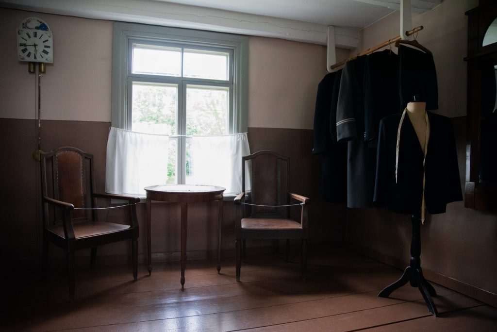 Räätälin tuvan vastaanottotila, jossa on kaksi tuolia, pöytä ja tanko, jolta riippuu valmiita pukuja.