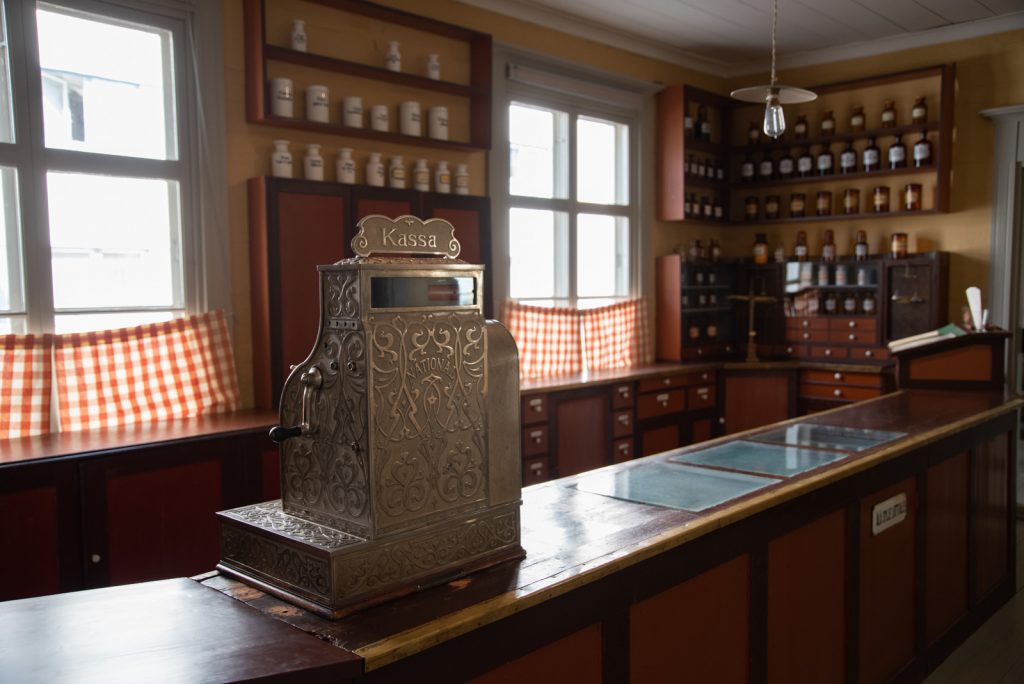 Apteekkimyymälän myyntitiski ja takana olevat hyllystöt sekä myyntitiskin päällä oleva koristeellinen kassakone.
